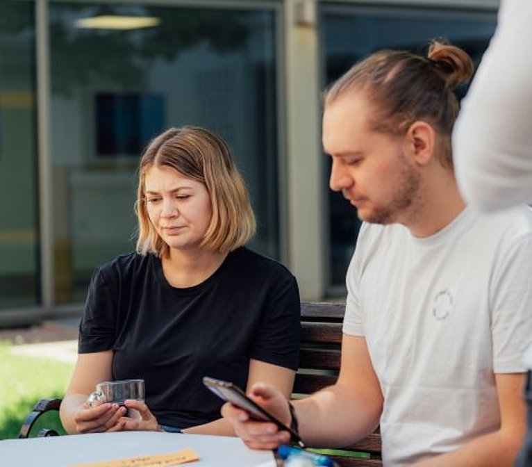Zwei Personen sitzen draußen am Tisch; eine hält ein Glas, der andere schaut aufs Smartphone.
